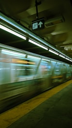 A subway train moves quickly through an underground station. Fluorescent lights illuminate the platform, and the train appears blurred due to its speed. A directional sign is visible overhead, and the yellow safety line runs parallel to the edge of the platform.
