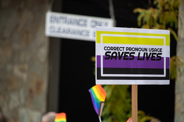 A sign held up with bold text stating 'Correct Pronoun Usage Saves Lives,' featuring a background with black, yellow, white, and purple colors. Nearby, there are small rainbow flags waving. The surroundings include some greenery and a blurred sign indicating an entrance.