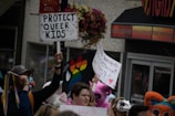 A crowd of people holds up various colorful signs supporting LGBTQ+ rights. One prominent sign reads 'Protect Queer Kids', while another bears a rainbow design. Participants are dressed vibrantly and some wear costumes, adding a festive and supportive atmosphere.