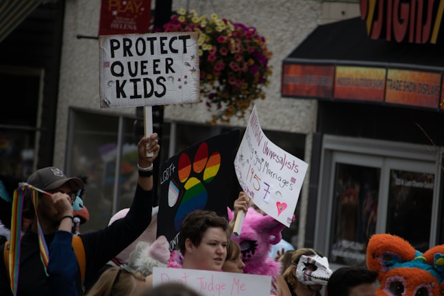 A crowd of people holds up various colorful signs supporting LGBTQ+ rights. One prominent sign reads 'Protect Queer Kids', while another bears a rainbow design. Participants are dressed vibrantly and some wear costumes, adding a festive and supportive atmosphere.