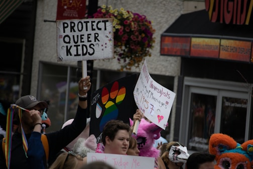 A crowd of people holds up various colorful signs supporting LGBTQ+ rights. One prominent sign reads 'Protect Queer Kids', while another bears a rainbow design. Participants are dressed vibrantly and some wear costumes, adding a festive and supportive atmosphere.