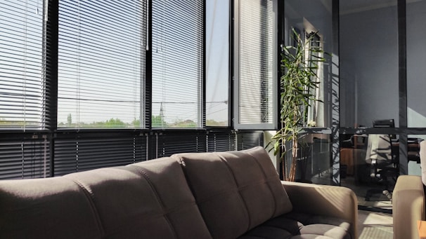 A cozy office or living area with large windows covered by horizontal blinds, allowing in filtered daylight. In the foreground, there is a plush brown sofa positioned under the windows. A tall green plant is placed near the windows, and in the background, there are pieces of office furniture including chairs and desks visible through the transparent partition.