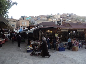 A bustling marketplace with various stalls selling goods. Vendors and customers are visible, with one person pushing a stroller. Traditional buildings in the background display satellite dishes and other modern amenities.