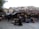 A bustling marketplace with various stalls selling goods. Vendors and customers are visible, with one person pushing a stroller. Traditional buildings in the background display satellite dishes and other modern amenities.