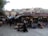 A bustling marketplace with various stalls selling goods. Vendors and customers are visible, with one person pushing a stroller. Traditional buildings in the background display satellite dishes and other modern amenities.