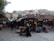 A bustling marketplace with various stalls selling goods. Vendors and customers are visible, with one person pushing a stroller. Traditional buildings in the background display satellite dishes and other modern amenities.