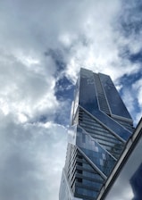 A modern skyscraper with a sleek, angular design rises towards a sky filled with clouds. The building's glass facade reflects the light, creating an interplay between the structure and the cloud-filled sky.