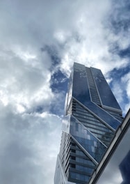 A modern skyscraper with a sleek, angular design rises towards a sky filled with clouds. The building's glass facade reflects the light, creating an interplay between the structure and the cloud-filled sky.
