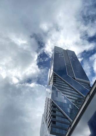 A modern skyscraper with a sleek, angular design rises towards a sky filled with clouds. The building's glass facade reflects the light, creating an interplay between the structure and the cloud-filled sky.