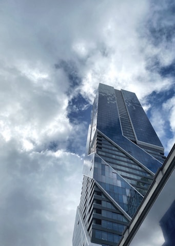 A modern skyscraper with a sleek, angular design rises towards a sky filled with clouds. The building's glass facade reflects the light, creating an interplay between the structure and the cloud-filled sky.