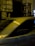 Posters are pasted on a textured stone wall at night, featuring a person's face with a contemplative expression. A blurred car passes by in the foreground, suggesting motion and speed. Bright lights and their reflections create additional visual interest.