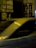 Posters are pasted on a textured stone wall at night, featuring a person's face with a contemplative expression. A blurred car passes by in the foreground, suggesting motion and speed. Bright lights and their reflections create additional visual interest.