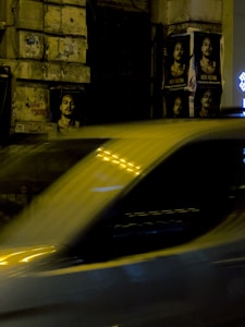 Posters are pasted on a textured stone wall at night, featuring a person's face with a contemplative expression. A blurred car passes by in the foreground, suggesting motion and speed. Bright lights and their reflections create additional visual interest.