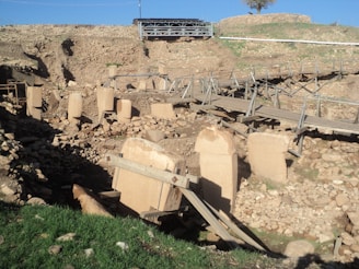 Ancient stone pillars stand within a partially excavated archaeological site surrounded by rubble and wooden walkways. The foreground features some grass, while the background shows an elevated metallic structure and remnants of stone walls.