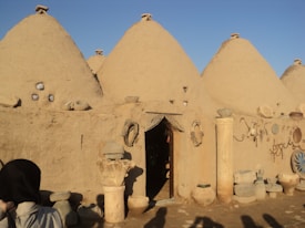 A series of traditional beehive-shaped mud brick houses with small openings and a wooden door. Various pots, stones, and decorative items are placed on and around the structures. The scene is bathed in warm sunlight, with a clear blue sky overhead.