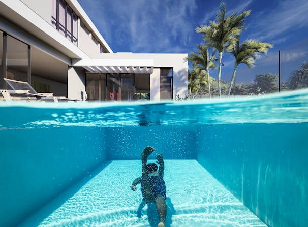 A luxurious modern house features an elegant outdoor swimming pool. The scene is split, showing both underwater and above the waterline. A person is swimming underwater, and palm trees can be seen alongside the contemporary architecture. The clear blue sky complements the serene and inviting atmosphere.
