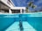 A luxurious modern house features an elegant outdoor swimming pool. The scene is split, showing both underwater and above the waterline. A person is swimming underwater, and palm trees can be seen alongside the contemporary architecture. The clear blue sky complements the serene and inviting atmosphere.