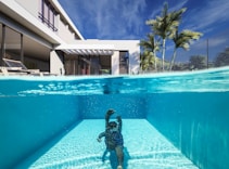 A luxurious modern house features an elegant outdoor swimming pool. The scene is split, showing both underwater and above the waterline. A person is swimming underwater, and palm trees can be seen alongside the contemporary architecture. The clear blue sky complements the serene and inviting atmosphere.