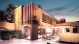 A modern two-story house with a sleek design features a large patio area with lounge chairs and a swimming pool. The exterior is predominantly glass with rectangular structures and soft, warm lighting creating a welcoming atmosphere. Palm trees and well-maintained hedges border the property, adding a tropical touch.