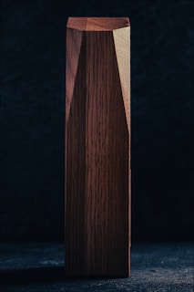 A wooden rectangular prism standing vertically against a dark background. The surface of the wood is smooth, showcasing the natural grain and rich brown color. Light highlights one side, emphasizing the texture and dimensions of the object.