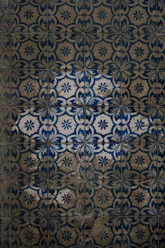 An intricate pattern of vintage tiles with an ornate floral design in shades of blue and beige. Some tiles are worn and have a weathered appearance, adding to the aged aesthetic.