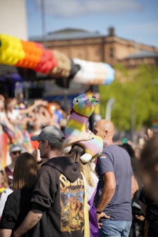 A festive outdoor event with a crowd of people, some wearing colorful attire and accessories. A person is holding a large, multicolored stuffed llama. In the background, there are bright decorations and a building partially visible. The atmosphere suggests celebration and joy.
