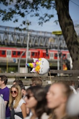A unicorn-shaped balloon with colorful patterns is held above a crowd of people. The scene includes several casually dressed individuals enjoying a sunny outdoor event, with a tree and a train in the background.