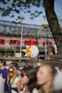 A unicorn-shaped balloon with colorful patterns is held above a crowd of people. The scene includes several casually dressed individuals enjoying a sunny outdoor event, with a tree and a train in the background.