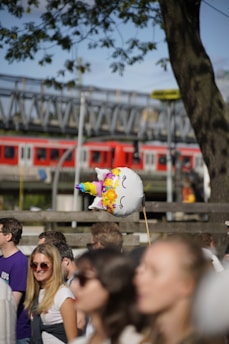 A unicorn-shaped balloon with colorful patterns is held above a crowd of people. The scene includes several casually dressed individuals enjoying a sunny outdoor event, with a tree and a train in the background.