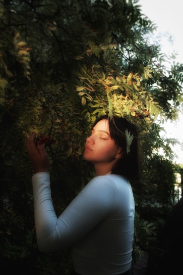 A person gently holding a branch with berries, surrounded by lush green foliage. Sunlight filters through the leaves, creating a serene and peaceful atmosphere. The individual appears calm and content, with closed eyes and a subtle smile.