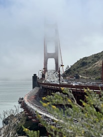 The iconic Golden Gate Bridge is partially shrouded in fog, with numerous vehicles traveling across it. The bridge's red metal structure contrasts with the green foliage in the foreground and the surrounding landscape featuring rolling hills.