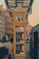 A bus stop sign labeled A1 with route maps displayed below. The sign and maps are encased in a yellow frame. People are gathered nearby, and a bus is visible in the background on a city street during the evening.