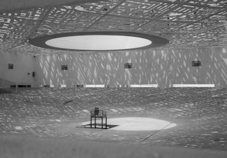 A spacious, modern architectural space with an intricate patterned ceiling casting complex shadows on the floor. Central to the room is a solitary chair placed in a spotlight. The walls are smooth and minimalist, contributing to the stark geometric aesthetics.