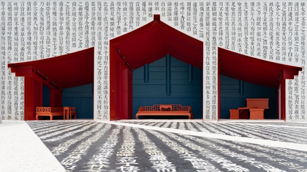 Three red-roofed wooden structures with traditional Asian design elements stand against a backdrop filled with columns of black Chinese characters. The floor also displays large white characters on a dark background, creating a dramatic visual contrast. The structures contain red benches and tables with intricate patterns.