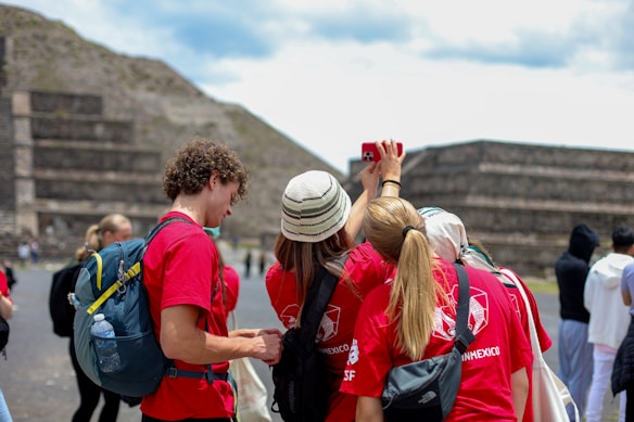 A group of people wearing red shirts with backpacks are taking a selfie in an outdoor setting with ancient stone structures in the background. One person is holding a phone while others gather around, smiling and joining in the photo.