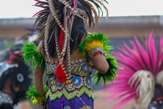 A person wearing colorful traditional attire adorned with vibrant feathers and intricate patterns. The clothing features bright green and yellow feathered arm decorations and a detailed pattern on the back. The scene is lively and filled with movement, with additional feathered costumes visible in the background.