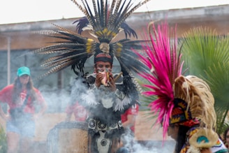 A person is dressed in traditional attire with a large feathered headdress, playing a conch shell amidst rising smoke. Another individual in a similar outfit is positioned in the foreground, partially visible. In the background, two people wearing casual clothing stand and observe.
