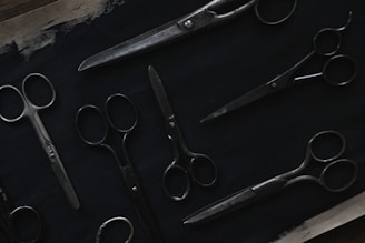 A variety of scissors are arranged on a dark surface. The scissors are of different sizes and styles, featuring metal blades and handles. The image is dimly lit, highlighting the metallic sheen against the dark background.
