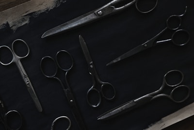A variety of scissors are arranged on a dark surface. The scissors are of different sizes and styles, featuring metal blades and handles. The image is dimly lit, highlighting the metallic sheen against the dark background.