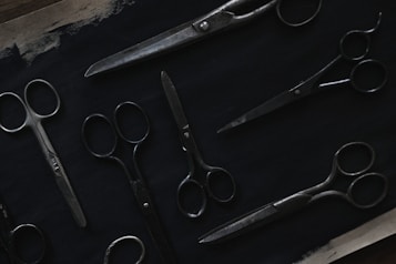 A variety of scissors are arranged on a dark surface. The scissors are of different sizes and styles, featuring metal blades and handles. The image is dimly lit, highlighting the metallic sheen against the dark background.