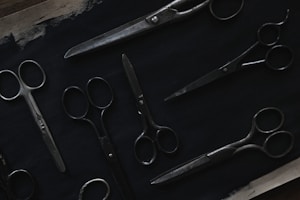A variety of scissors are arranged on a dark surface. The scissors are of different sizes and styles, featuring metal blades and handles. The image is dimly lit, highlighting the metallic sheen against the dark background.