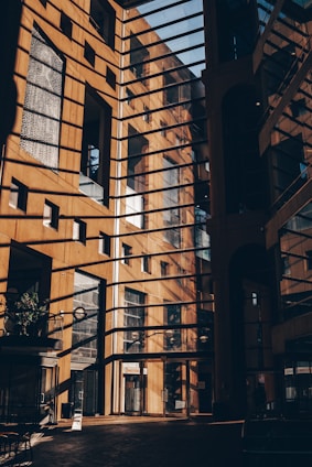 A modern architectural structure with large glass windows and metal frames allowing sunlight to cast intricate shadows on the walls and floor. The interior showcases a sophisticated design with a warm color palette created by the sunlight.