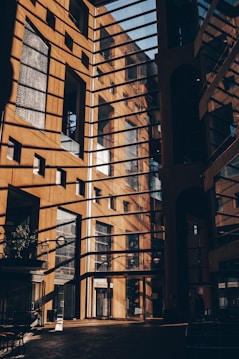 A modern architectural structure with large glass windows and metal frames allowing sunlight to cast intricate shadows on the walls and floor. The interior showcases a sophisticated design with a warm color palette created by the sunlight.