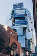 A modern, glass-fronted office building with the Deloitte logo at the top. The structure features an angular and unique architectural design with reflective blue-tinted windows. In the foreground, there is another building labeled ‘Centre,’ which contrasts with its traditional brown brick facade. The city environment is visible with some greenery at the edges.