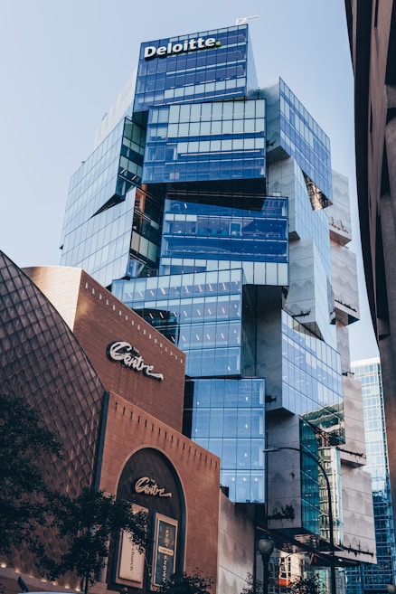 A modern, glass-fronted office building with the Deloitte logo at the top. The structure features an angular and unique architectural design with reflective blue-tinted windows. In the foreground, there is another building labeled ‘Centre,’ which contrasts with its traditional brown brick facade. The city environment is visible with some greenery at the edges.