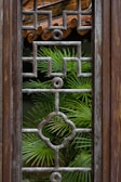 A wooden window frame features an intricate geometric grille design with a verdant palm plant visible through its lattice structure. The background shows part of a rustic tiled roof, contributing to the natural and earthy ambiance.