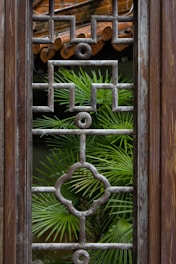 A wooden window frame features an intricate geometric grille design with a verdant palm plant visible through its lattice structure. The background shows part of a rustic tiled roof, contributing to the natural and earthy ambiance.