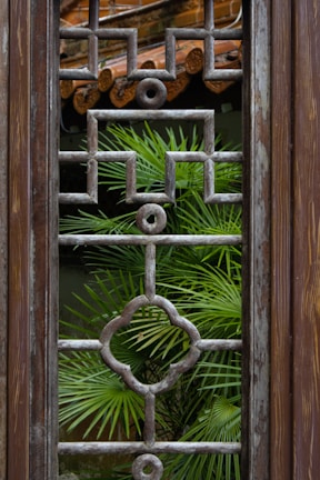 A wooden window frame features an intricate geometric grille design with a verdant palm plant visible through its lattice structure. The background shows part of a rustic tiled roof, contributing to the natural and earthy ambiance.