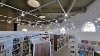 A spacious library with modern architecture features multiple rows of bookshelves filled with various books. The ceiling is high with unique circular designs and numerous pendant lights hanging above. Large arched windows allow natural light to enter, creating a bright and inviting atmosphere.