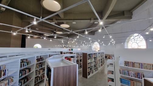 A spacious library with modern architecture features multiple rows of bookshelves filled with various books. The ceiling is high with unique circular designs and numerous pendant lights hanging above. Large arched windows allow natural light to enter, creating a bright and inviting atmosphere.